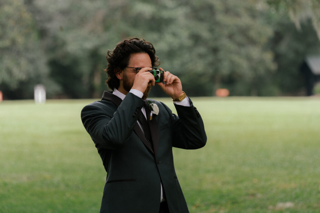 Groom in a dark green suit taking a photo with a disposable film camera during his wedding day in Nashville.